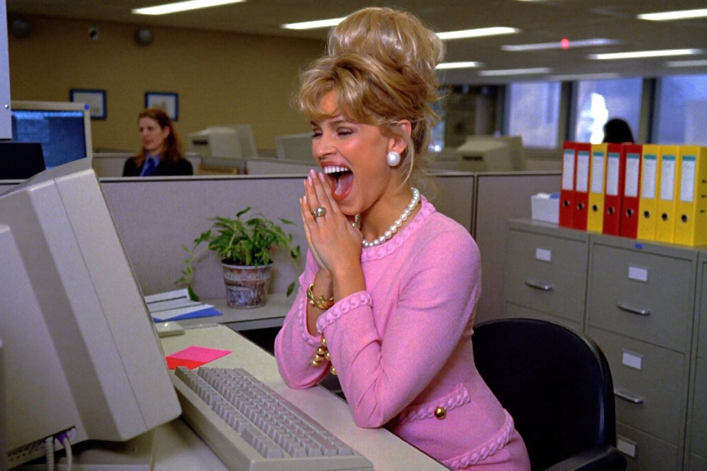 A woman in a pink suit in a 1990s office clasping her hands in delight at what has appeared on her computer screen